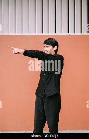 Un giovane uomo concentrato vestito con una camicia nera e pantaloni si staglia di profilo, puntando saldamente il dito indice verso destra contro una semplice parete arancione b Foto Stock