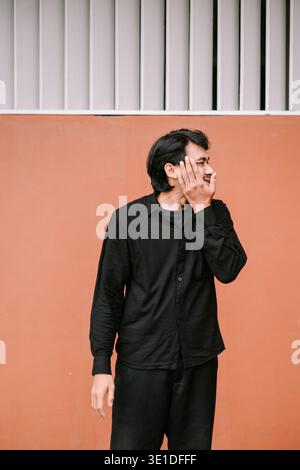 Un giovane asiatico con una camicia nera casual sorride timidamente mentre si copre la bocca contro una parete arancione. Sembra che sia divertito o stia cercando di sopprimere Foto Stock