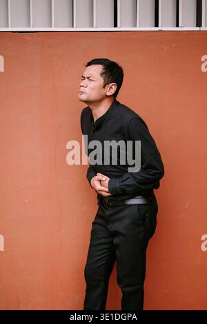 Un uomo che soffre di uno stomaco turbato, che tiene l'addome nel dolore. Si trova contro un muro arancione, mostrando disagio e malattia. Ideale per la salute Foto Stock