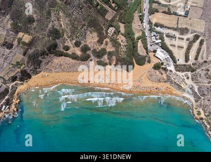 Vista dall'alto verso il basso della spiaggia di Ramla a Nadur, Gozo, con sabbia arancione, ombrelloni, mare turchese, surf line, strada costiera e parcheggio. Foto Stock