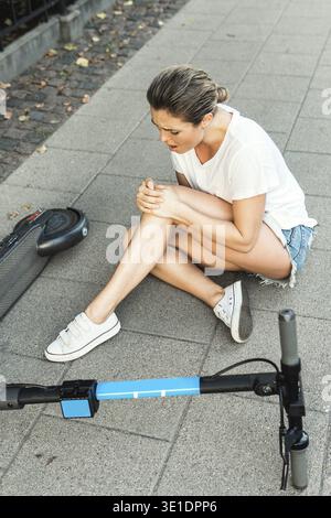 Giovane donna che soffre di dolore al ginocchio dopo un incidente di guida e-scooter su una strada della città Foto Stock