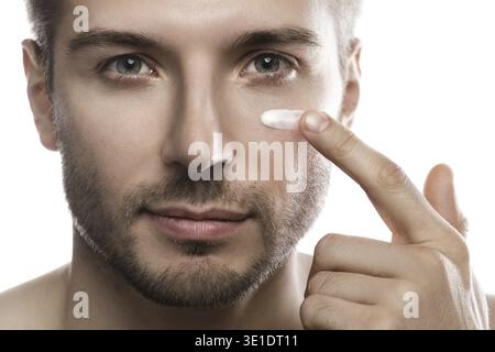 Bellezza maschile. Il giovane uomo sta applicando crema idratante e anti invecchiamento sul suo viso su sfondo bianco Foto Stock