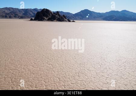 Un vasto piano di fango incrinato si estende verso un gruppo di formazioni rocciose scure sotto un cielo azzurro. Il paesaggio appare arido e desolato, con d Foto Stock