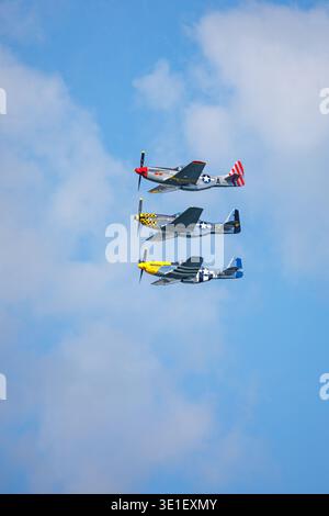 Tre aerei da caccia P-51 Mustang volano in formazione contro un cielo blu con nuvole sparse. I piani sono dipinti in varie livree storiche, in Foto Stock
