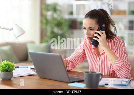 Lavoratore autonomo serio che parla al telefono controllando il computer portatile che lavora a casa Foto Stock