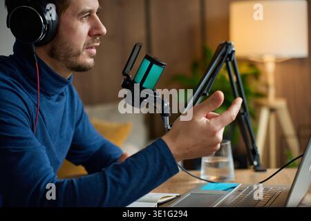 L'uomo che indossa le cuffie parla nel microfono del desktop vicino al computer portatile in un'area di lavoro di casa. Concetto di podcasting, streaming, comunicazione online e conten Foto Stock