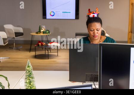 Donna afroamericana che lavora su due monitor in ufficio con fascia per renne e spazio fotocopie Foto Stock