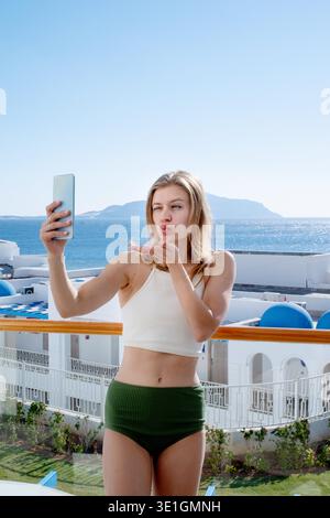 Vacanze e viaggi. Donna felice in costume da bagno che scatta selfie su un telefono cellulare in piedi sul balcone della camera d'albergo, vista mare || modello rilasciato Foto Stock