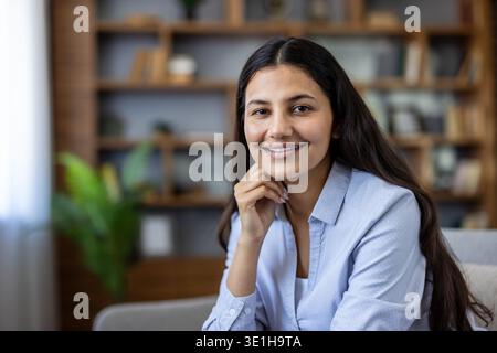 Giovane donna indiana adulta sorridente. Guarda la fotocamera con fiducia e professionalità. Seduti in un interno domestico con sfondo da libreria. Rappresenta il lavoro da casa e lo stile casual elegante Foto Stock