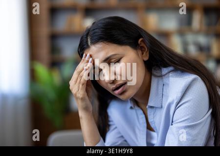 Giovane donna che sente un intenso mal di testa e dolore, toccando la fronte a occhi chiusi, simboleggiando angoscia, affaticamento e stanchezza mentale dovuti a pressioni schiaccianti e lotte quotidiane Foto Stock