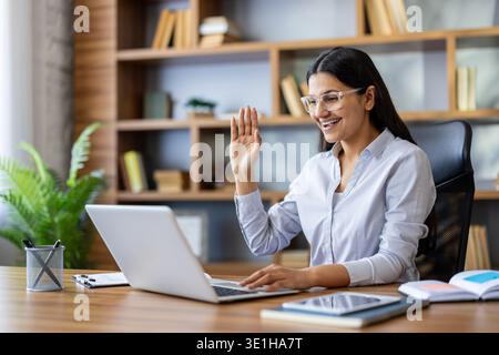 Donna che indossa gli occhiali sorridenti, che si muove a mano mentre partecipa a una riunione virtuale su un laptop dal suo ufficio domestico, si gode il lavoro remoto e la comunicazione online Foto Stock