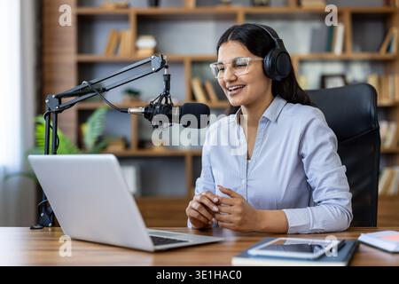 Giovane donna che indossa cuffie e occhiali, che parla con un microfono professionale a una scrivania con un laptop, creando contenuti audio digitali per la sua trasmissione online Foto Stock