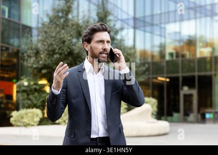 Uomo d'affari con un abito in piedi di fronte a un moderno edificio di uffici in vetro, che parla su uno smartphone e gesta con la mano durante una conversazione all'aperto Foto Stock