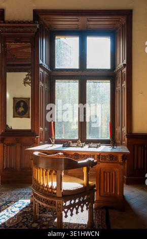 Studio storico tradizionale caratterizzato da elaborati mobili in legno intagliato che includono una scrivania ornata e una sedia collocata davanti ad una finestra a bovindo. Legno pregiato pa Foto Stock