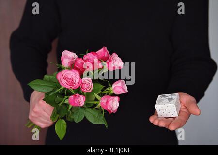 Un uomo con abiti neri ha un mazzo di rose rosa brillante e una piccola confezione regalo. Foto di alta qualità Foto Stock