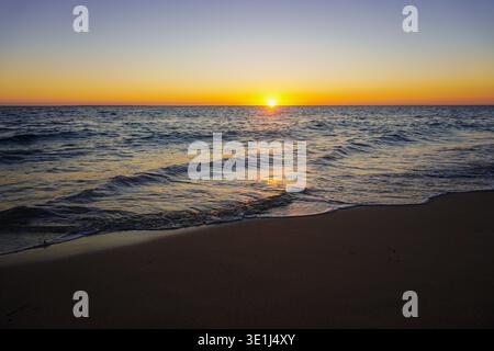 Il sole tramonta sull'orizzonte dell'oceano con piccole onde che si innalzano sulla spiaggia sabbiosa sotto il cielo limpido Foto Stock
