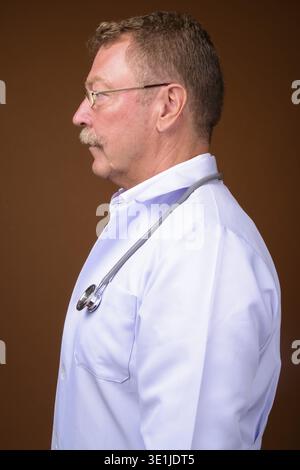 Foto in studio di un bellissimo medico anziano con baffi che indossano occhiali da vista su sfondo marrone Foto Stock