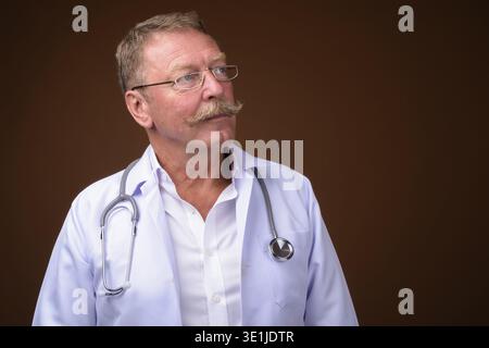 Foto in studio di un bellissimo medico anziano con baffi che indossano occhiali da vista su sfondo marrone Foto Stock