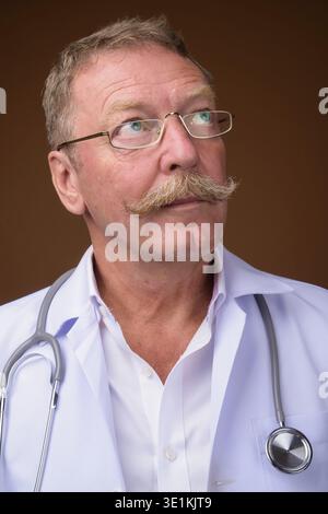 Foto in studio di un bellissimo medico anziano con baffi che indossano occhiali da vista su sfondo marrone Foto Stock