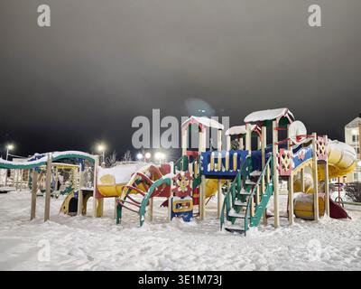 Parco giochi per bambini coperto di neve bianca durante la notte. Le colorate strutture di gioco sono visibili sotto le luminose luci esterne. Foto Stock