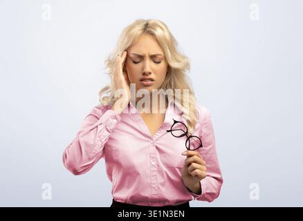 La donna tiene gli occhiali e mostra disagio nella testa mentre indossa una camicia rosa Foto Stock