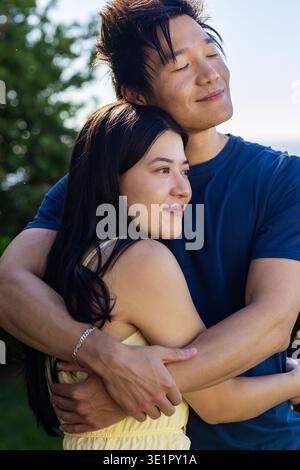 Coppia asiatica che abbraccia, uomo che indossa una maglietta blu che tiene la donna in abito giallo, braccialetto d'argento nel parco Foto Stock