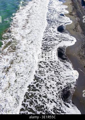 Le onde si infrangono sulla spiaggia sabbiosa nel pomeriggio. La marea arriva, creando schiuma sulla costa mentre il sole splende sull'acqua. Foto Stock