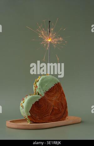 Festività natalizie candela sparkler anniversario anniversario celebrazione a forma di cuore pistacchio croissant su sfondo verde con spazio fotocopie. Pasta sfoglia nuova pasticceria dolce alla moda. Esperimento di impasto cremoso Foto Stock