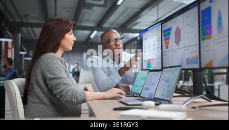 Giovane analista di business afroamericano e collega che esamina i grafici dei dati finanziari, discutendo i KPI Foto Stock