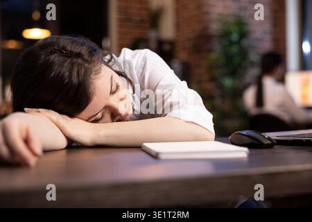 La manager aziendale si prende un breve pisolino al suo tavolo durante il turno di notte. Una donna d'affari stanca si addormenta alla scrivania dell'ufficio, sopraffatta da lunghe ore lavorative in un ambiente di lavoro moderno. Foto Stock