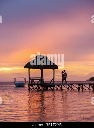 Il tramonto mozzafiato illumina il cielo mentre una coppia condivide un momento tenero su un molo di legno a Mauritius. La tranquillità dell'acqua cattura l'essenza del romanticismo e dei viaggi di lusso. Foto Stock