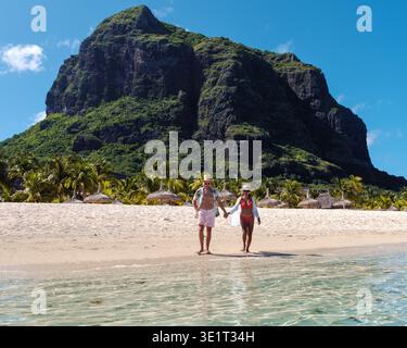 Una coppia gioiosa cammina mano in mano lungo le rive di una splendida spiaggia a Mauritius, circondata da vegetazione lussureggiante e maestose scogliere. Le Morne Mauritius Foto Stock