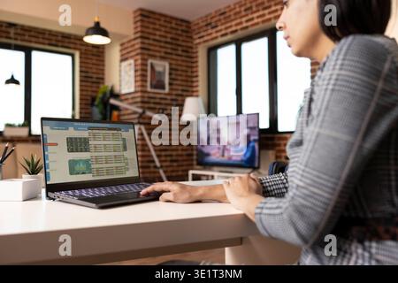 Una donna incentrata lavora da casa, utilizzando un laptop per analizzare le statistiche aziendali e monitorare le spese. Donna d'affari indiana che esamina le tendenze finanziarie negli appartamenti murati. Foto Stock
