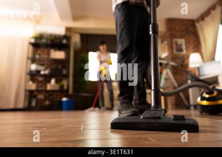 Pulizia individuale maschile per pavimenti in legno con aspirapolvere mentre la donna pulisce sullo sfondo. Primo piano dell'uomo che ripulisce il soggiorno, mantenendo la pulizia in un appartamento moderno durante le faccende domestiche. Foto Stock