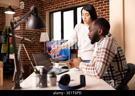 Fidanzata di supporto che parla di software su notebook con uno sviluppatore maschile concentrato alla scrivania dell'ufficio domestico. Coppia multietnica che lavora in remoto insieme e brainstorming sullo sviluppo di app per un'azienda tecnologica. Foto Stock