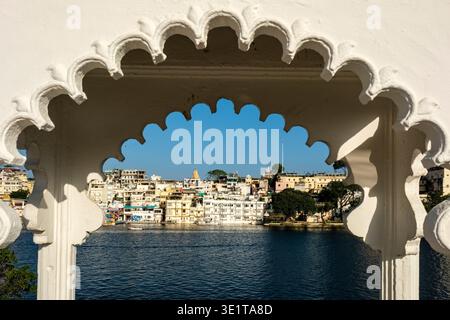 Gli edifici bianchi di Udaipur e la guglia del Tempio Jagdish sono visti attraverso l'arco decorativo in pietra di un padiglione sul lago Pichola. Foto Stock