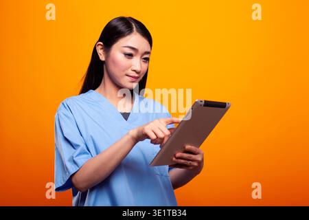 Infermiera donna che tiene il tablet e controlla le cartelle cliniche online. Giovane donna asiatica vestita in uniforme medica, che lavora con un dispositivo digitale, isolato su sfondo giallo, focalizzato sulla salute. Foto Stock