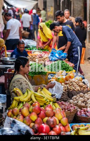 Una donna in un mercato locale di Udaipur seleziona i prodotti da una bancarella piena di mele, melograni, banane e verdure varie. Foto Stock