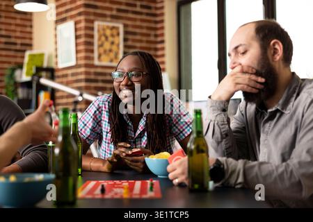 Allegra donna afro-americana con gli occhiali, sorridente mentre tiene in mano le carte da gioco, preparandosi al suo turno. È seduta con gli amici intorno al tavolo, pronta a godersi una vivace sessione di gioco al chiuso. Foto Stock