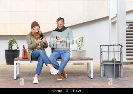 Coppia senior seduta su una panchina di legno nella piazza all'aperto, smartphone a scorrimento con borsa marrone Foto Stock