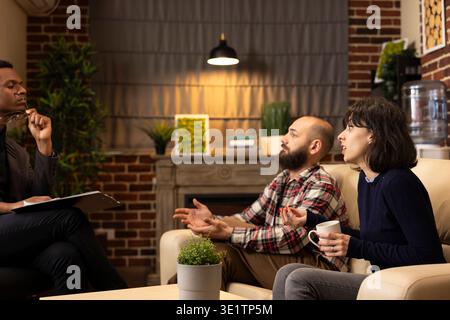Lo psichiatra si siede con gli appunti al ginocchio mentre la coppia caucasica discute i conflitti di relazione sul divano. Il terapista ascolta attentamente, analizzando i problemi di matrimonio e fornendo una guida professionale. Foto Stock