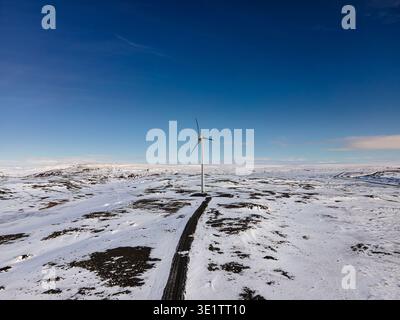 La vista aerea di una turbina eolica solitaria è una sentinella tra il paesaggio arido e innevato, un faro di energia pulita sotto il vasto cielo blu, l'Islanda. Foto Stock