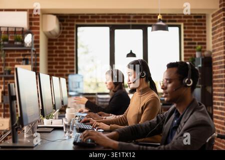 Attenzione selettiva alla donna asiatica che indossa una cuffia, che lavora con colleghi diversi e utilizza il computer in uno spazio di lavoro con pareti a mattoni. I dipendenti multietnici lavorano alla propria scrivania, parlando con i clienti. Foto Stock