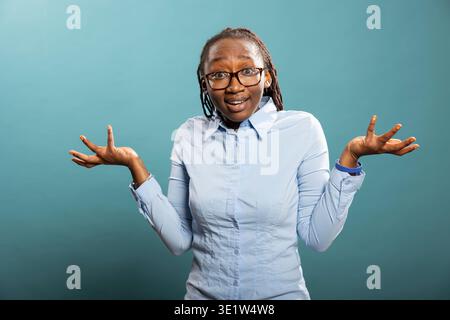 Ritratto in studio di donna afroamericana con espressione confusa, che solleva spalle e mani in un gesto interrogativo. Modello femminile nero che appare sconcertato, in piedi su sfondo blu. Foto Stock