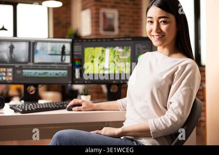 Donna asiatica della fotografia sorridente con fiducia nello studio di casa seduto su due monitor. Creative editor femminile che utilizza un software di classificazione dei colori per produrre video cinematografici in remoto. Foto Stock