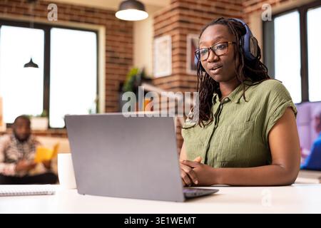 Donna afroamericana che utilizza un computer portatile e le cuffie, partecipa a un workshop aziendale online da casa. Una donna imprenditrice partecipa a sessioni di apprendimento interattive, sedute alla scrivania durante una riunione remota con i fornitori Foto Stock