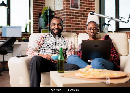 Ritratto dell'uomo che guarda la televisione e della donna che usa il laptop, che naviga in internet e che si diverte con il film in tv. Mangiare un pasto fast food dalla consegna da asporto e bere bottiglie di birra. Foto Stock