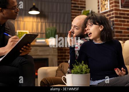Lo psicologo afroamericano osserva la coppia caucasica seduta a schiena durante la sessione di consulenza. La donna condivide apertamente le sfide di relazione mentre il terapeuta maschile nero prende appunti con attenzione. Foto Stock