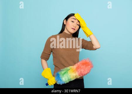 Domestica asiatica esausta in guanti di gomma gialli che contengono piume, mostrando un'espressione stanca dopo una lunga giornata di lavoro. Schiacciata donna che sfrega la fronte e si sente affaticata. Foto Stock