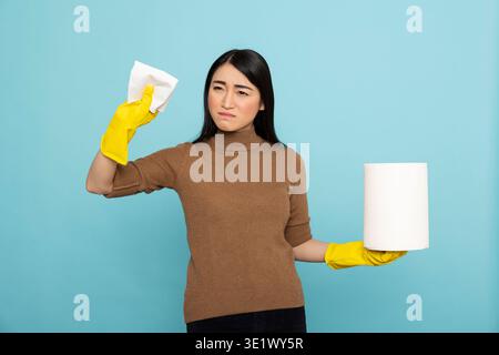 Giovane donna asiatica concentrata in guanti di gomma gialli con asciugamano usa e getta, preparata ad affrontare le attività di pulizia. Governante serio pronto a garantire igiene e ordine a casa. Foto Stock